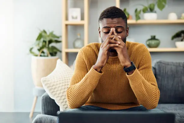 Man sits on a sofa with elbows on knees, hands clasped over his face, looking down at a laptop in front of him, appearing stressed or thoughtful.