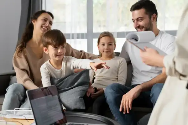 Family of four sits together on a couch, smiling as a presenter shows papers; a laptop is open on a nearby table—likely a family meeting or counseling session.